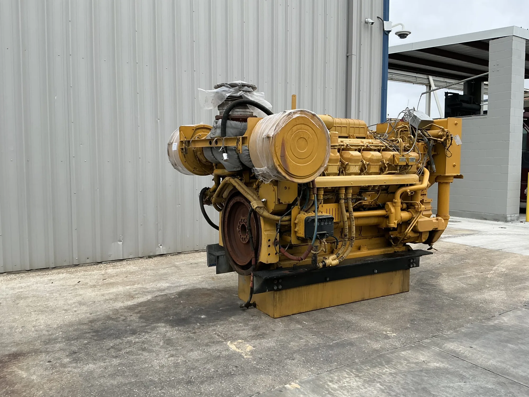 Diesel generator set in yellow, featuring a large circular front cover, with hoses and wiring visible, outdoors beside a gray corrugated metal wall