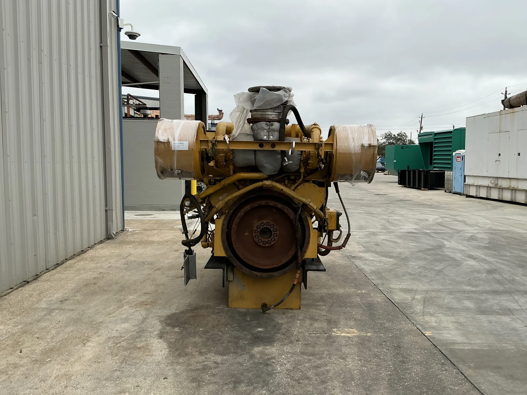 Large yellow industrial diesel engine with a prominent front flywheel, exposed hoses, and components wrapped in plastic